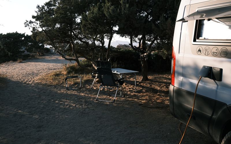 Kastenwagen am Stellplatz unter Kiefern, davor Tisch und Campingstühle